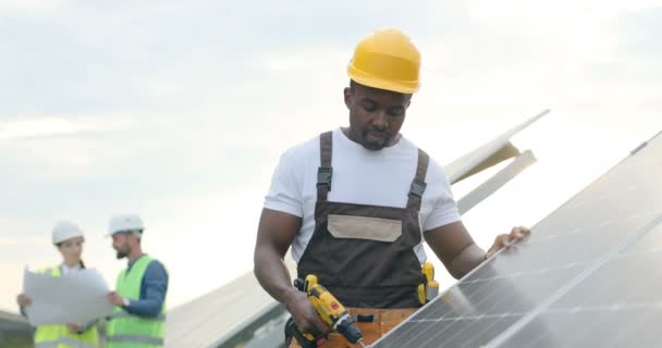 Portret młodego Afroamerykanina mechanika w żółtym hełmie wykonującego otwory w panelu słonecznym z wiertłem elektrycznym. — Wideo stockowe