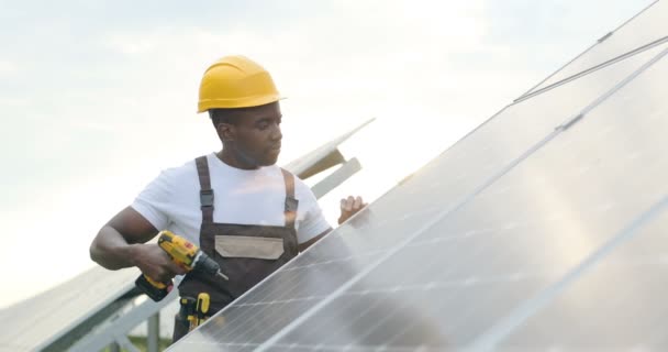 Bonito jovem afro-americano mecânico em capacete amarelo fazendo furos no painel solar com broca elétrica. Movimento lento. — Vídeo de Stock