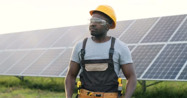Hermoso trabajador afroamericano de pie en medio de vidrio con energía solar y sonrisas. Hombre en casco protector con los brazos cruzados posa para la cámara. — Vídeos de Stock