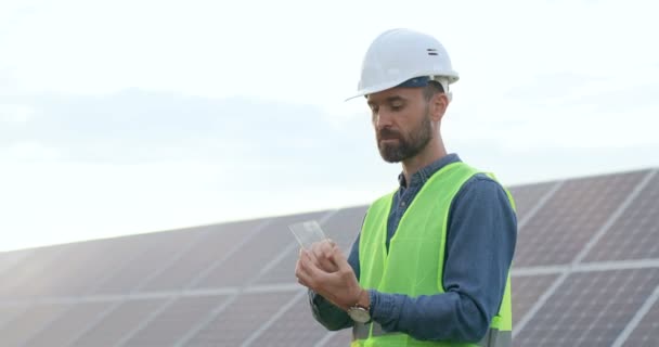 Hezký kavkazský technik stojící venku mezi solárními panely a zkoumá složené tablety. Práce na slunné moderní farmě. — Stock video