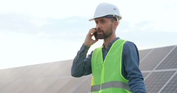 Ein hübscher kaukasischer Techniker mit weißem Schutzhelm steht zwischen Sonnenkollektoren und telefoniert rege mit seinen Händen.. — Stockvideo