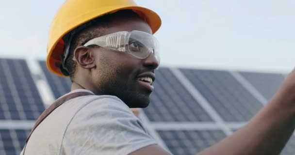 Zbliżenie portret młodego, przystojnego, afrykańskiego mężczyzny w twardej czapce i goglach mówiącego o energii słonecznej i bateriach. — Wideo stockowe