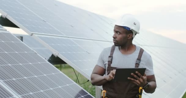 Bonito afro-americano mecânico masculino em pé fora perto de painéis solares segurando tablet. Homem a montar uma rede eléctrica alternativa. — Vídeo de Stock