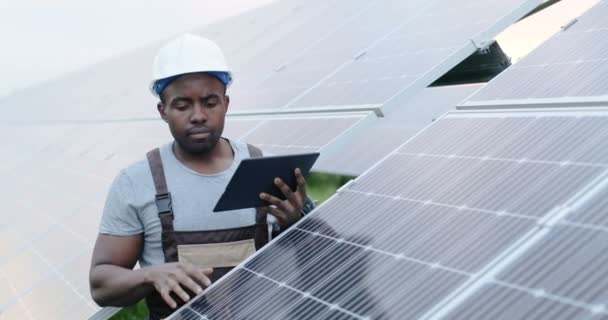 Όμορφος αφροαμερικανός μηχανικός στέκεται έξω κοντά σε εναλλακτικό εργοστάσιο παραγωγής ενέργειας κρατώντας tablet στα χέρια. Επαγγελματικός έλεγχος λειτουργίας ηλιακών συλλεκτών. — Αρχείο Βίντεο