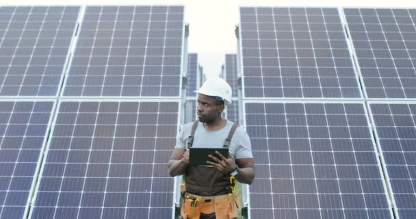 Guapo afroamericano mecánico masculino en uniforme de pie delante de filas de paneles solares sosteniendo tableta digital y mirando hacia otro lado. — Vídeo de stock