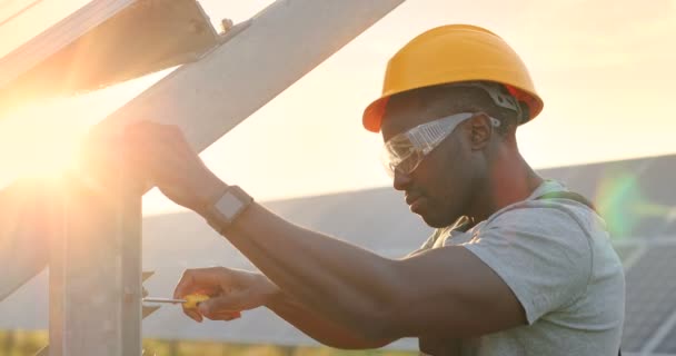 Close up portrait of african american mechanic in yellow hard hat working with screwdriver on solar panel fixing frame. — Stock Video