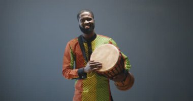 Geleneksel kıyafetler giymiş neşeli Afro-Amerikan bir gencin portresi. Partiye hoş geldin der gibi davul çalıyor. Davullu yakışıklı erkek davulcu mutlu gülümsüyor. Afrika 'da çalan müzisyen.