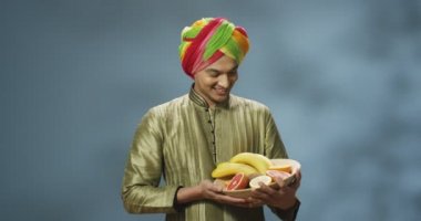 Genç Hindu yakışıklı bir adamın portresi. Tardışık kıyafetler içinde ve kafasında sarık var. Kameraya neşeyle gülümsüyor ve ellerinde meyvelerle tabağı tutuyor. Hindistan konseptine hoş geldiniz. Meyveli mutlu adam..