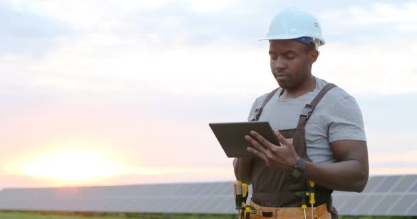 Dışarıda elektronik tablet üzerinde çalışan üniformalı ve koruyucu kasklı Afrikalı Amerikalı tamircinin portresi. Alternatif enerji santrali kavramı. — Stok video
