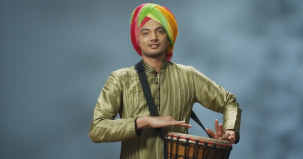 Retrato de un músico indio en turbante tocando el tambor y sonriendo alegremente a la cámara. Hombre guapo en traje tradicional jugando en bongo con la cara feliz. Músico con instrumento musical. — Vídeos de Stock