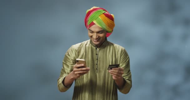 스마트폰으로 터번 쇼핑하면서 행복하게 웃는 즐거움을 주는 젊은 힌두 청년. 인터넷을 통해 돈을 벌고 신용 카드로 지불하는 세련 된 인도 남자. 행복 한 남성 고객 전화를 통해 구매. — 비디오
