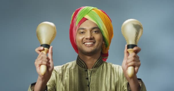 Retrato de un joven hindú guapo con turbante sonriendo alegremente y jugando en maracas. Primer plano del chico feliz de la India sosteniendo instrumentos musicales y haciendo rima a la música mientras se sacude. — Vídeos de Stock