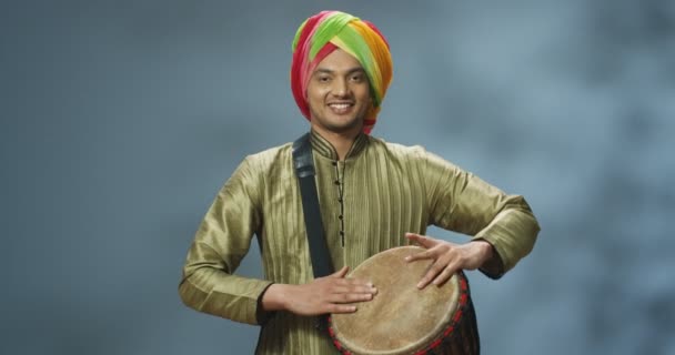 Retrato de músico indiano masculino em turbante tocando no tambor e sorrindo alegremente para a câmera. Homem bonito em roupas tradicionais brincando no bongo com rosto feliz. Músico com instrumento musical. — Vídeo de Stock