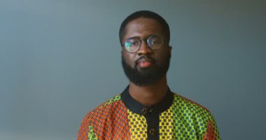 Çekici Afro-Amerikalı gözlüklü genç bir adamın gülümseyerek kameraya bakışının portresi. Renkli gömlekli, neşeli, yakışıklı bir erkek, gri duvarda mutlu bir şekilde gülümsüyor..
