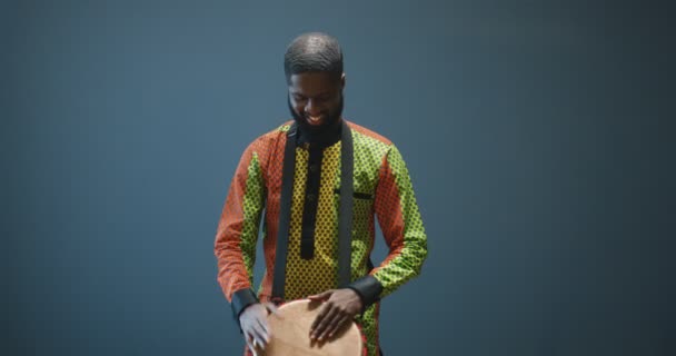 Porträt eines afroamerikanischen Musikers mit Brille, der auf die Trommel klopft und in die Kamera lächelt. Schöner Mann in traditioneller Kleidung, der mit glücklichem Gesicht auf Bongo spielt. Musiker mit Musikinstrument. — Stockvideo