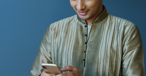 Close up de jovem hindu bonito homem alegre em roupas tradicionais sorrindo, tocando no smartphone e mensagem de texto. Bonito cara indiano digitando sms no celular. Conversando nas redes sociais. — Vídeo de Stock