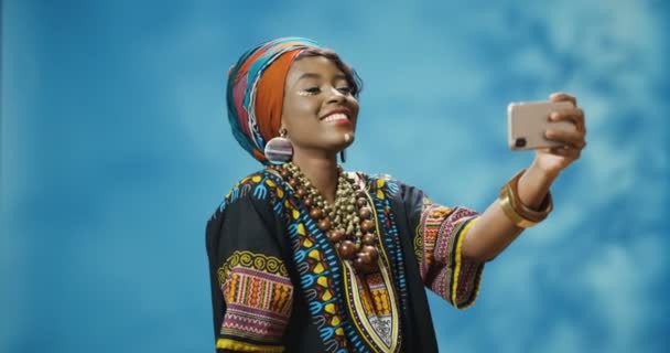 Stijlvolle Afro-Amerikaanse jonge mooie vrouw in traditionele kleding glimlachen vrolijk en poseren voor smartphone camera en het nemen van selfie foto 's. Mooie vrouw maakt foto 's met telefoon. — Stockvideo