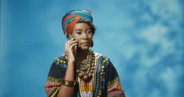 Jonge mooie Afro-Amerikaanse vrouw in traditionele kleding die vrolijk praat op de mobiele telefoon en glimlacht. Aantrekkelijke vrolijke vrouw spreekt op mobiele telefoon. Meisjespraat aan de telefoon met glimlach. — Stockvideo