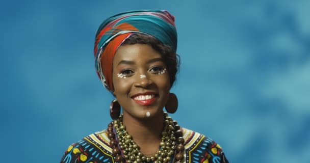 Портрет афроамериканской красивой молодой женщины в красочном традиционном наряде, смотрящей в камеру и радостно улыбающейся. Довольно счастливая женщина радостно улыбается стоя на синем фоне стены. — стоковое видео