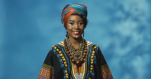 Portrétní snímek atraktivní mladé Afroameričanky v tradičním oblečení dívající se s úsměvem do kamery. Radostná krásná fena v barevné košili šťastně se usmívá na modrém pozadí zdi. — Stock video