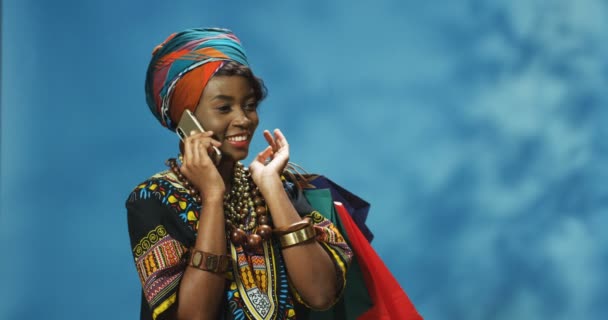 Vrolijke jonge Afro-Amerikaanse vrouw in tulband vrolijk pratend op mobiele telefoon, lachend en pakketjes over de schouder vasthoudend na het winkelen. mooi meisje spreken op mobiele telefoon met glimlach en tassen. — Stockvideo