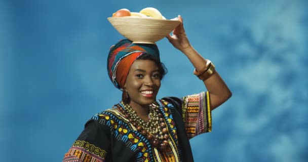 Portret van jonge vrolijke glimlachte mooie Afro-Amerikaanse vrouw met bord met tropisch fruit op het hoofd en kijkend naar de camera. Aantrekkelijk meisje tonen vruchten en glimlachen gelukkig. — Stockvideo