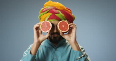 Renkli türbanlı Hindu genç yakışıklı adamın iki parça greyfurtla gözlerini kapayıp açtığı yakın plan. Meyveli çekici bir adam. Geleneksel kıyafetlerle portresi yapılmış..