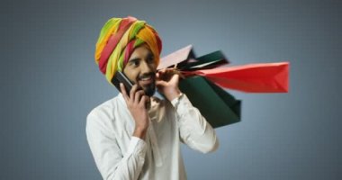 Geleneksel türbanlı yakışıklı genç Hindu adam alışveriş yaptıktan ve cep telefonuyla konuştuktan sonra sırt üstü paketleri tutuyor. Çantalı mutlu Hintli erkek cep telefonuyla konuşuyor..