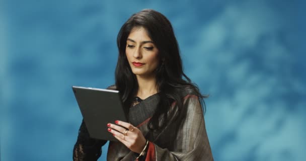 Elegante attraente indù giovane donna allegra in abito tradizionale toccando e scorrendo sul dispositivo tablet. Bella ragazza digitando e messaggiando sul computer tablet con sorriso su sfondo blu. — Video Stock