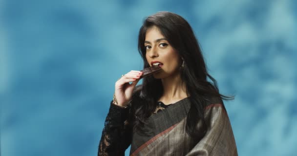 年轻美丽的印度教女人穿着传统服装，咬着一块黑巧克力，笑着。漂亮的印度时尚女孩，吃着糖果，品尝着美味. — 图库视频影像