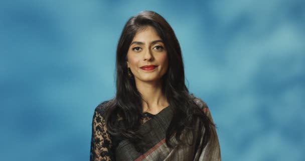 Portrait prise de vue de la jeune femme heureuse joyeuse élégante hindoue dans la tenue traditionnelle de l'Inde souriant à la caméra sur fond bleu. Gros plan de charmante femme aux lèvres rouges et au sourire. — Video