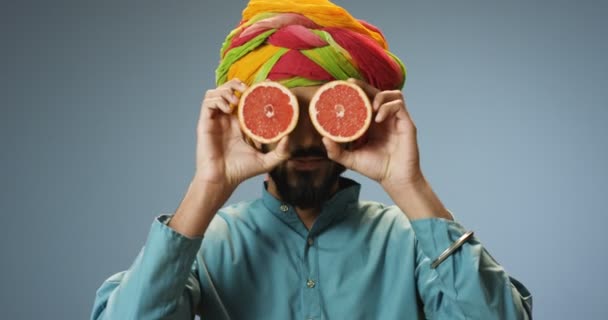 Close up de alegre jovem hindu bonito homem em turbante colorido segurando duas metades de toranja fechando os olhos e abrindo. Atraente cara feliz com frutas em mãos retrato em roupas tradicionais — Vídeo de Stock