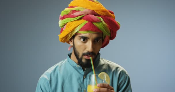 カラフルなターバンで陽気な若いハンサムなヒンズー教徒の男が藁と笑顔でオレンジジュースを飲んで閉じます。幸せな笑顔男の肖像画上の新鮮な飲み物をすすります灰色の壁の背景. — ストック動画