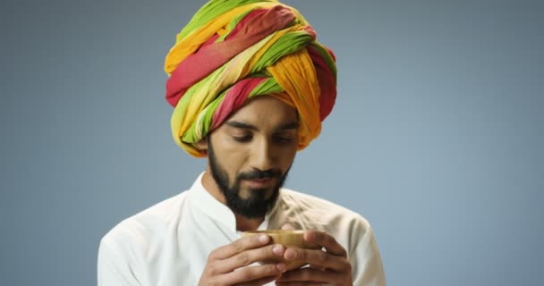 Primo piano di bel giovane indù in turbante colorato tradizionale e con barba sorseggiando bevanda calda dalla tazza. Attraente indiano maschio bere tè su sfondo grigio parete. — Video Stock