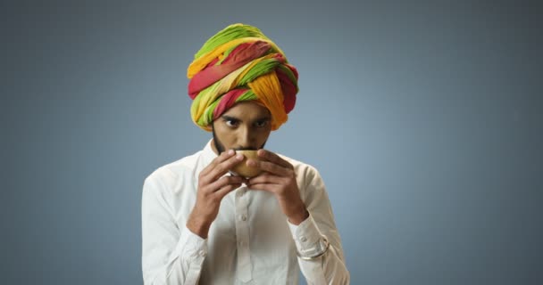 伝統的なカラフルなターバンでハンサムな若いヒンズー教徒の男と髭カップから熱い飲み物をすすります。魅力的なインドの男性はお茶を飲み、灰色の壁の背景に笑顔. — ストック動画
