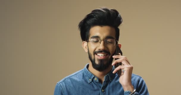 Homem hindu bonito novo com barba e em óculos falando alegremente no telefone celular e sorrindo. Homem indiano feliz atraente falando no celular. cara em jeans camisa falar no telefone com sorriso. — Vídeo de Stock
