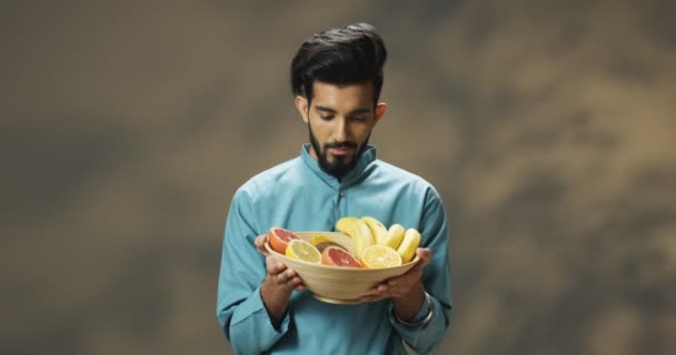年轻而快乐的英俊的印度教男子的画像，手拿着热带水果展示盘，看着镜头。迷人的男人展示水果，开心的笑着. — 图库视频影像