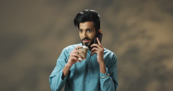 Χαρούμενος νεαρός όμορφος Ινδός με μπλε πουκάμισο να μιλάει στο κινητό και να πίνει ζεστό ποτό. Χαρούμενος χαμογελαστός τύπος που μιλάει στο κινητό και πίνει καφέ ή τσάι. — Αρχείο Βίντεο