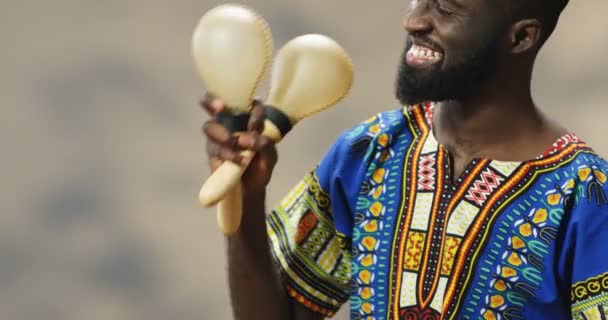 Bonito jovem afro-americano em roupas tradicionais sorrindo alegremente e brincando em maracas. Fechar-se de cara feliz segurando instrumentos musicais e fazendo ritmos à música enquanto treme. — Vídeo de Stock