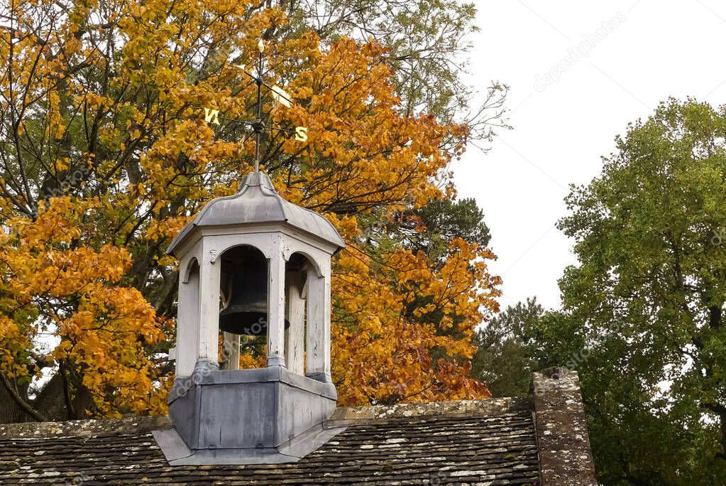 Autumnal image of a roof mounted Victorian bell tower, the structure with weathered, peeling white paint. Gilt metal weathervane. Background of fall coloured leaves on trees. Space for text.