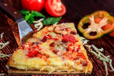 Koyu ahşap desenli lezzetli pizza dilimi taze sebzeler ve fırında salam, jambon, baharatlı sosis dolmalık biber ve mozzarella peyniri.