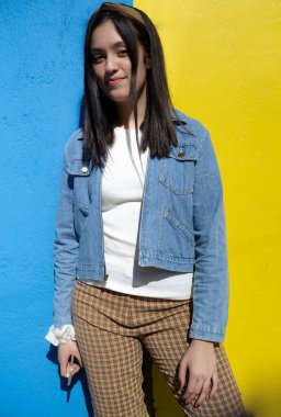 Gülümseyen ve günlük kıyafetler giyen genç bir kız mavi ve sarı arka planlı kameraya bakıyor.