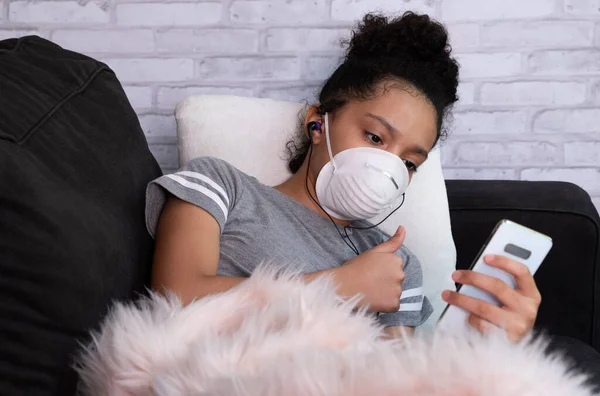 Female child wearing protective mask,  using mobile phone at sofa, show thumb up sign during Video conference with mobile phone. corona virus concept.Video call.