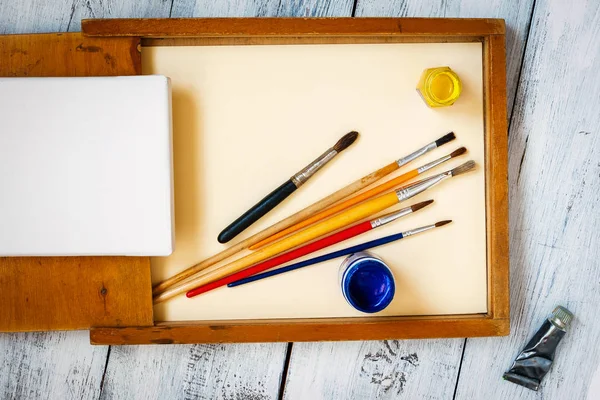 Brushes, colored paints in jars and a stretcher with a white canvas stand in a wooden box