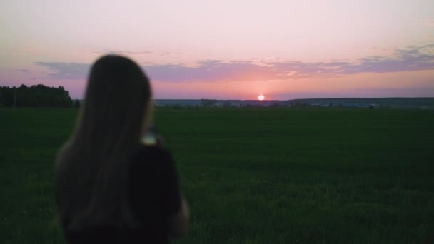 Σιλουέτα ενός κοριτσιού που φωτογραφίζει το ηλιοβασίλεμα στο τηλέφωνο στο χωράφι — Αρχείο Βίντεο