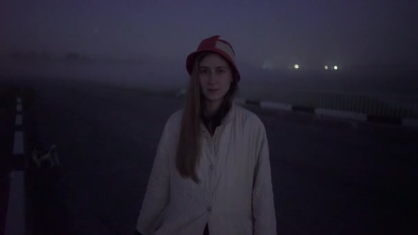 Žena v červenobílém klobouku a bílé bundě stojí v mlze a dívá se do kamery — Stock video