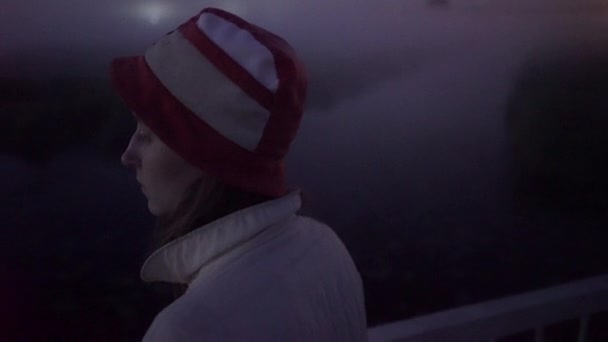 Menina bonita em chapéu vermelho-branco olhando para o nevoeiro junto ao rio à noite — Vídeo de Stock