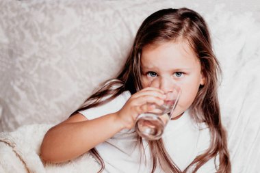 Hasta bir çocuk, beyaz bir yastıkla yatakta oturan bir kız, bardaktan su içen.