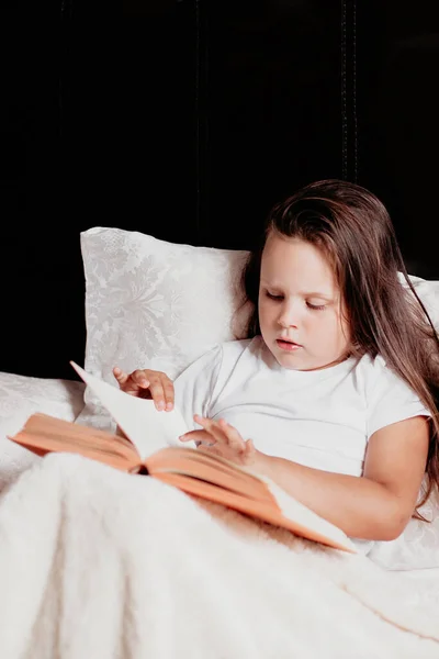 Enfant est assis sur le lit recouvert d'une couverture légère et d'un livre de lecture qu'il tient sur ses genoux, tournant la page avec sa main — Photo