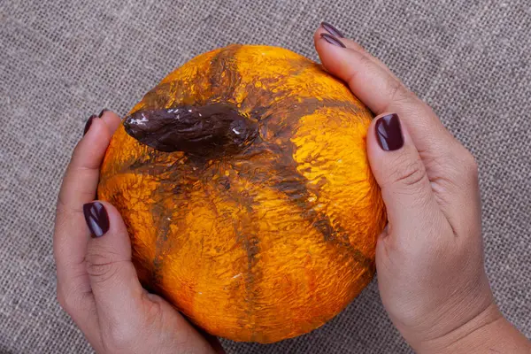 Zwei Hände halten einen hausgemachten orangefarbenen Pappmaché-Kürbis für Halloween auf einem Klempner-Hintergrund, Ansicht von oben — Stockfoto